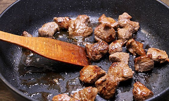 Обжаренные кусочки мяса в сковороде с деревянной лопаточкой