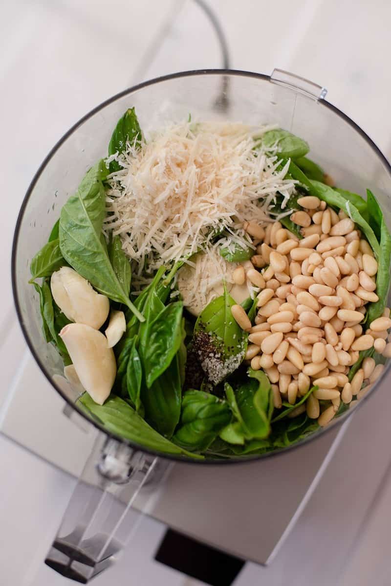 Overhead view of Pesto Sauce ingredients in the food processor, including grated parmesan cheese, pine nuts, basil, and garlic.