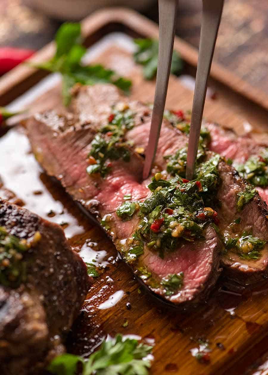 Slices of medium rare cooked steak on a wooden cutting board with Chimichurri Sauce